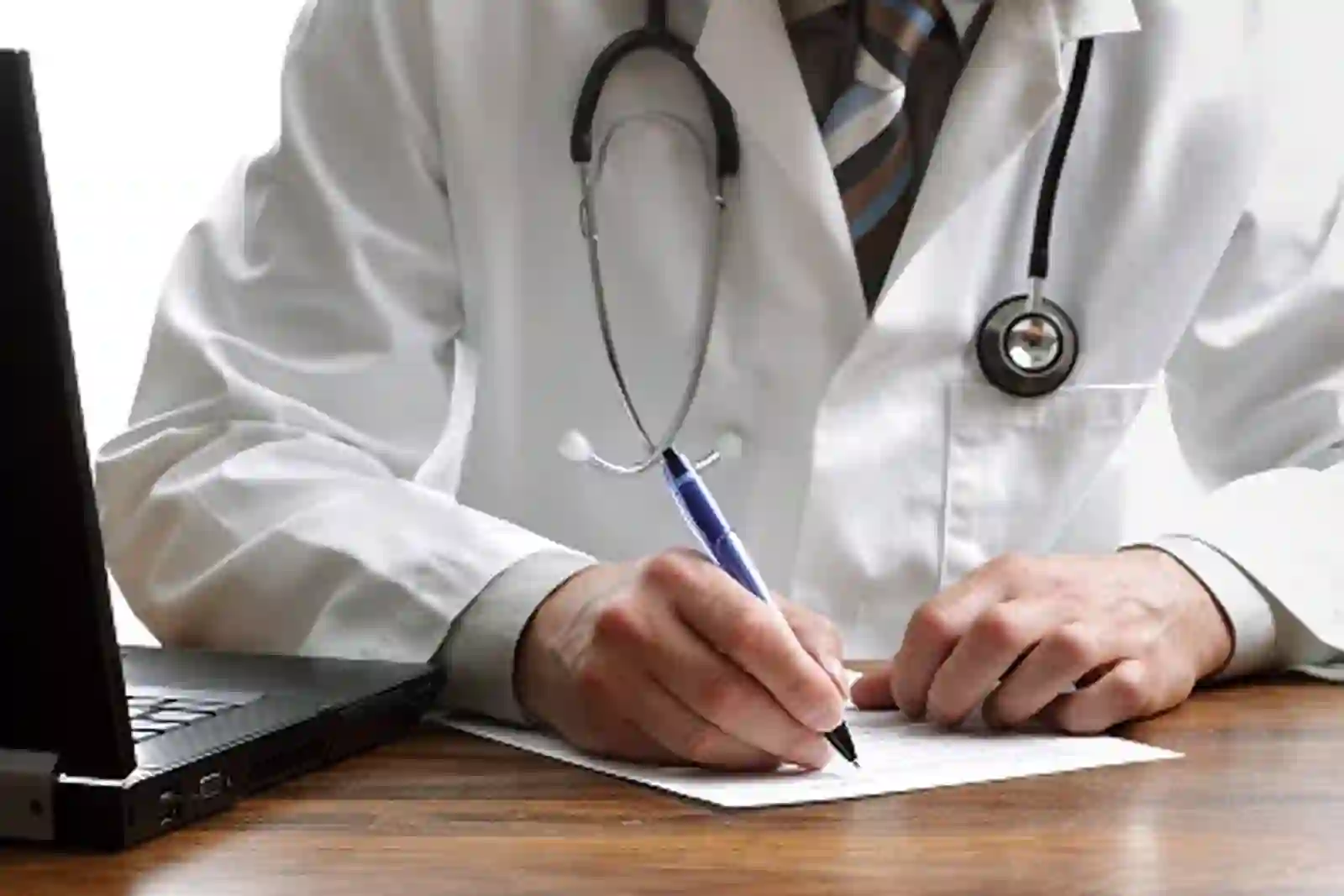 Doctor in a white coat writing a referral letter beside a laptop